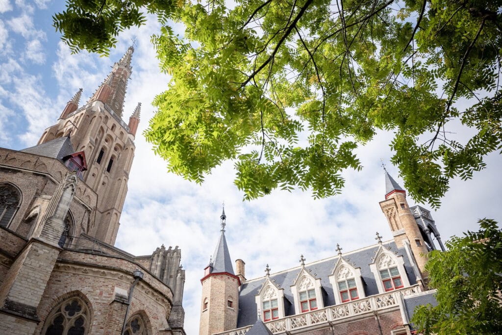 A view of a tall church tower with intricate design elements, surrounded by lush green leaves, under a partly cloudy sky.