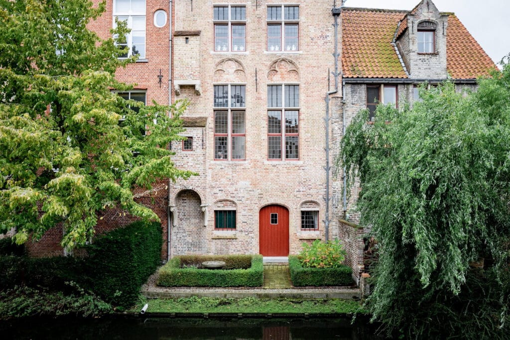 A brick building with large windows, a red door, and decorative stonework is surrounded by greenery and a canal. A manicured garden with low hedges is visible in front of the building.