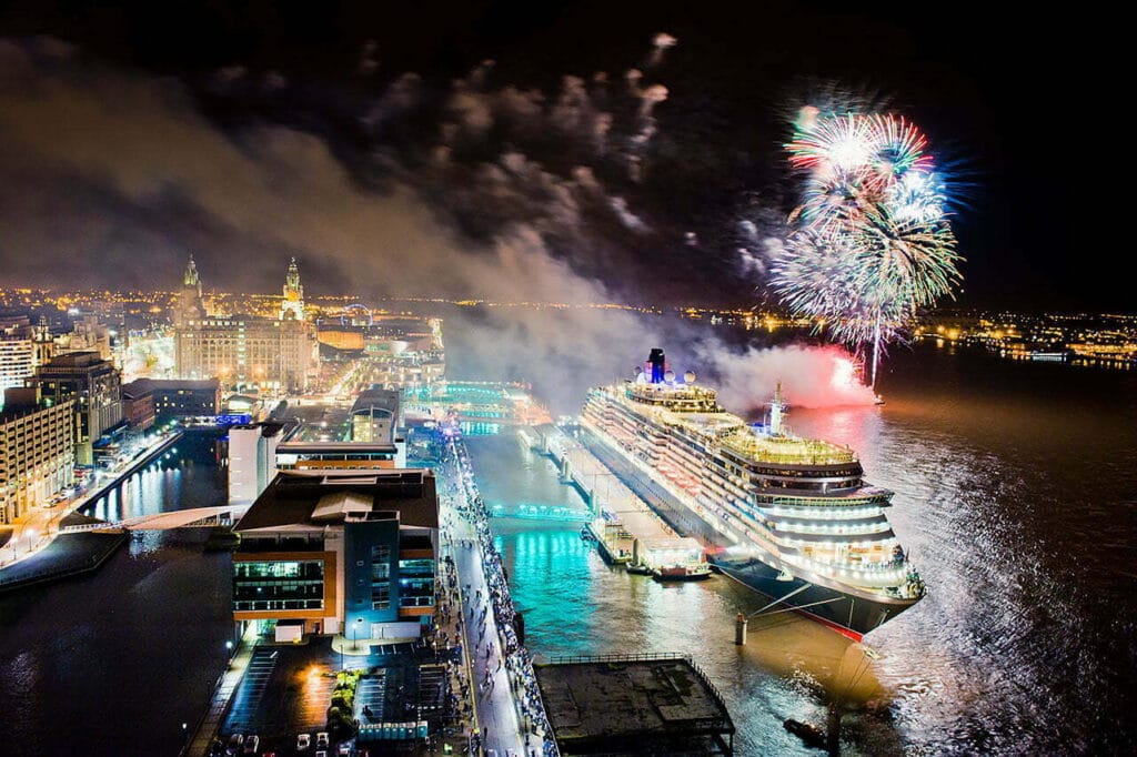 A nighttime aerial view of Liverpool waterfront featuring illuminated buildings, a large ship docked in the harbour, and vibrant fireworks lighting up the sky.
