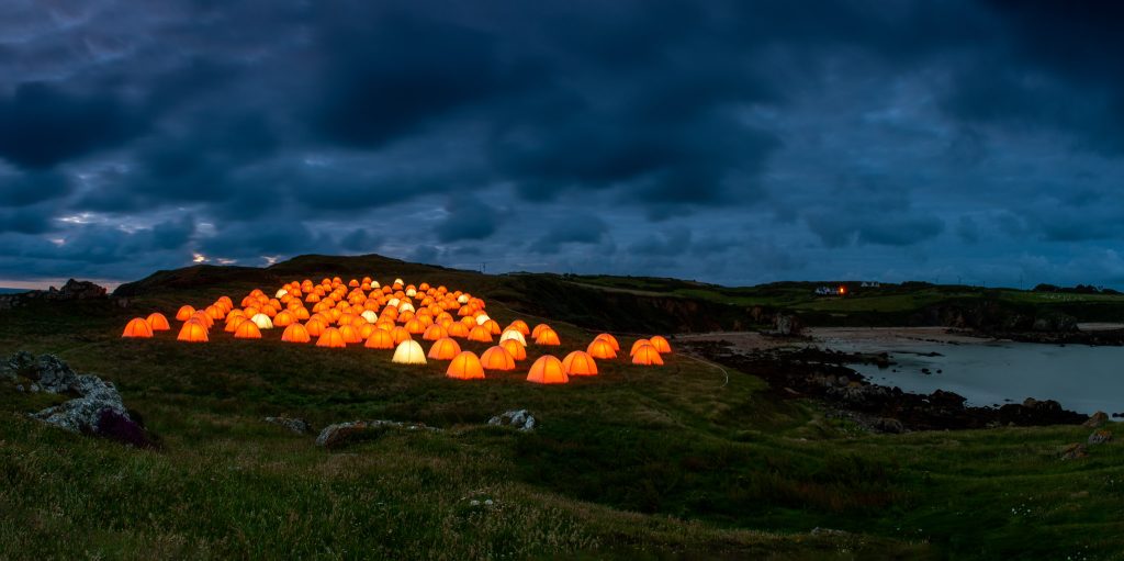 At dawn on top of a cliff on the edge of a bay, orange tents glow against the blue dusk sky. On the right hand side the tide is low in the bay.