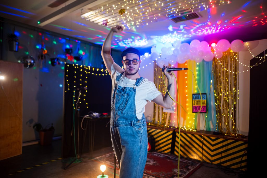 Person wearing dungarees holds a microphone and dances to an audience. In the background is a lit up rainbow wall with a sign saying "Queer joy is a protest."