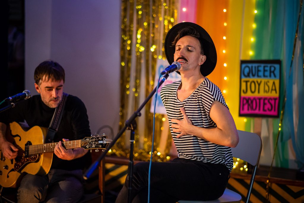 A person wearing a black and white striped t-shirt sits and sings to an audience at a gig in a cafe. Behind them is a sign saying "Queer joy is a protest."