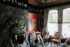 In a large room there is a 6 feet high lion coming out of the fireplace. There are papier-mâché body parts on chairs.