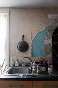 An old kitchen sink with various tins next to it. The wallpaper is torn and there is a drawing of a heart with an arrow through it.
