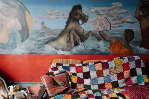 A painting of a hose in the ocean is painted directly onto a red wall. There is a couch with a multicoloured quilt on it.