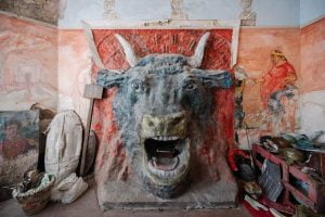 The head of a bull extends out from what would have been a fireplace. On the walls either side are faded paintings. The floor is covered by random artworks.