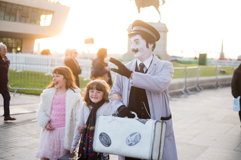 A man dressed as a luggage attendant with a white painted face performs for kids.
