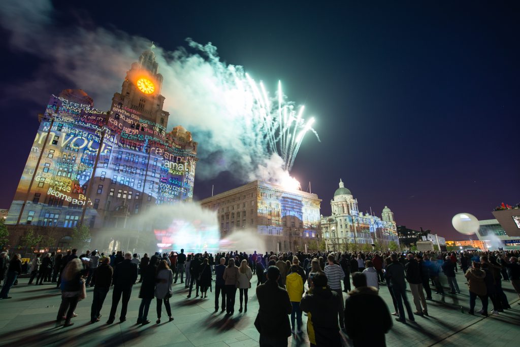 Fireworks shooting off the top of the Cunard building in the middle of the 3 Graces. On the left the Liver Building and on the right the Port of Liverpool building. A crowd watches projections on each.
