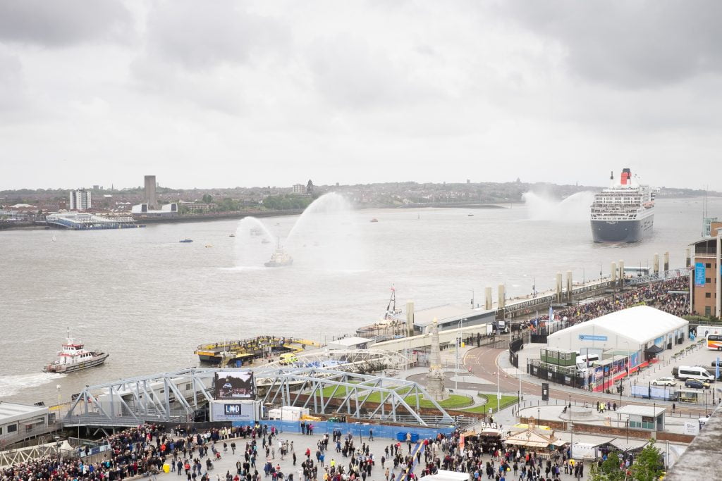 As a Cunard ship sails down the River Mersey, tugs spray jets of water into the air.