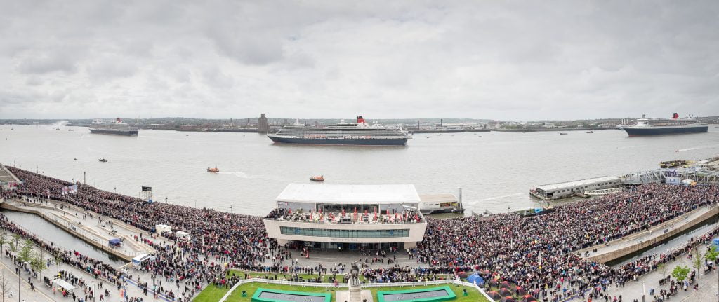 3 large cruise ships sailing down the River Mersey with a large crowd watching from the waterfront.
