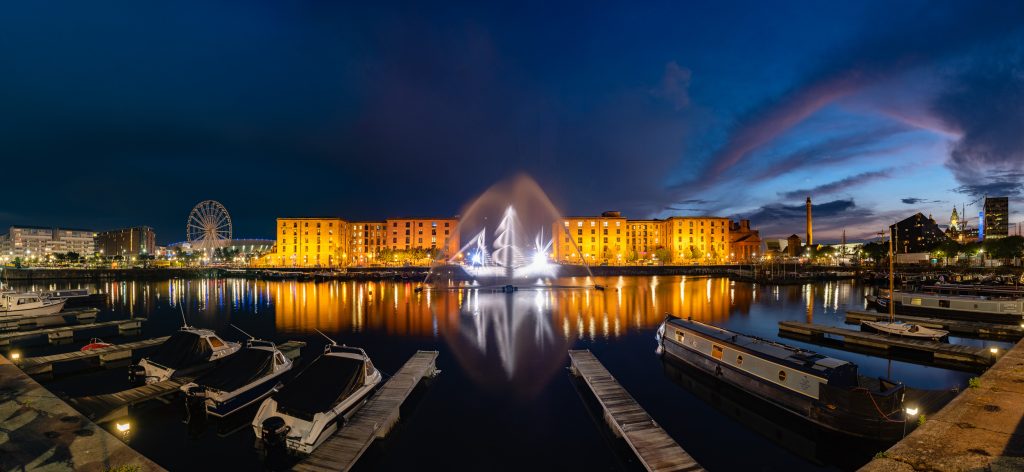 A ghostly vision of an old sailing ship is projected onto jets of water shooting up out of a dock. There are canal barges in front of the ghost ship. On the left is the arena and big wheel. On the right, with a bit of pink sunset in the sky, are the 3 Graces.