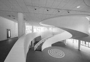 A modern, white interior featuring a sweeping staircase and curved walls, with large windows providing natural light. A spiral design is painted on the floor.