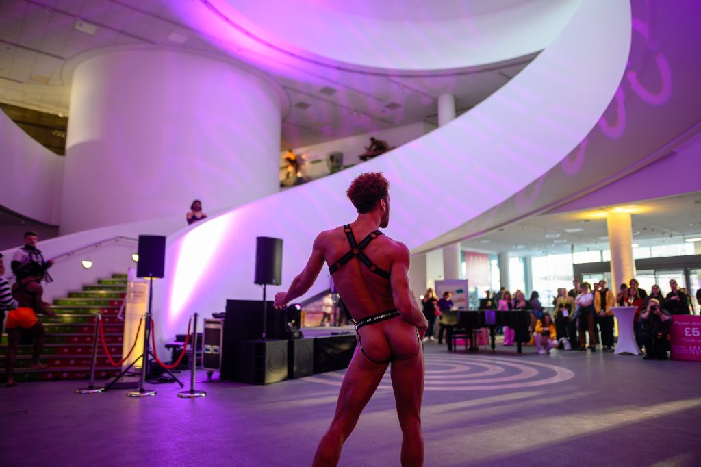 A man wearing a backless thong vogues inside a museum.