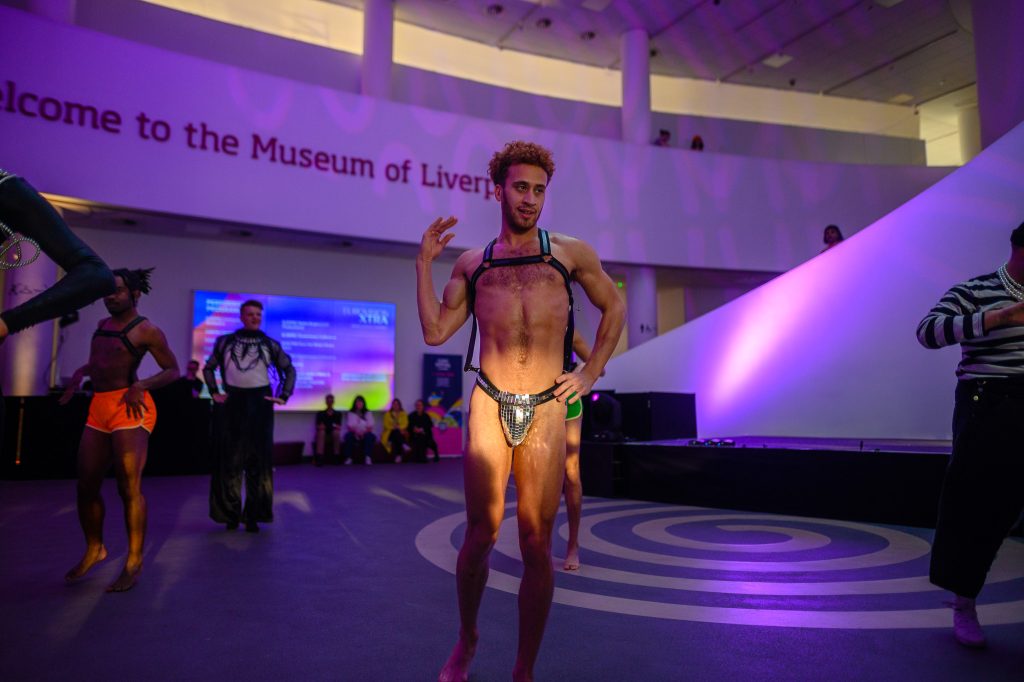 A man wearing only a disco ball styled thong vogues for the audience in the museum.