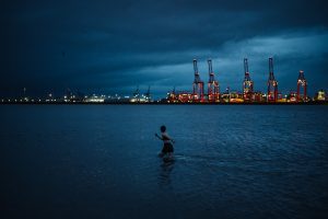 A woman runs in the waters of the River Mersey at dawn. On the horizon are tall red cranes waiting for a container ship.
