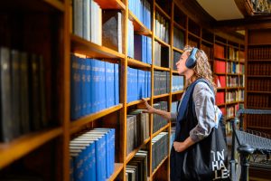 A woman wearing headphones listens to an audio tour while looking at various books in a library.