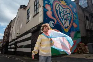 A man holds a transgender flag that is backlit by the sun outside a mural saying "Liverpool Loves You".