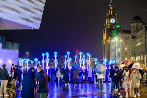 People gather around blue and pink balls on tall poles that are flashing and making music on the Liverpool watefrfont.