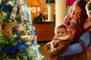 A black woman holds their smiling baby up to look at a maritme themed Christmas Tree.