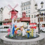 Colourful inflatables are positioned over an air vent outside the Moulin Rouge. They appear to be dancing with their air.