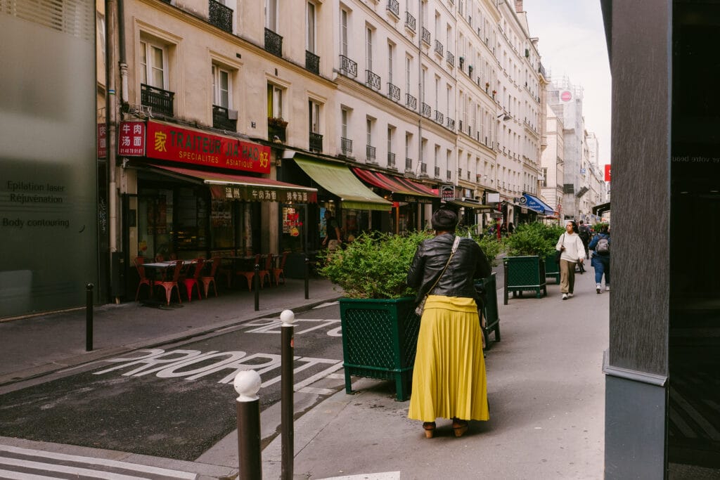 A woman, with her back to the camera, wears a yellow skirt and black coat. On the left of the street is a Chinese resturant with red stylings.