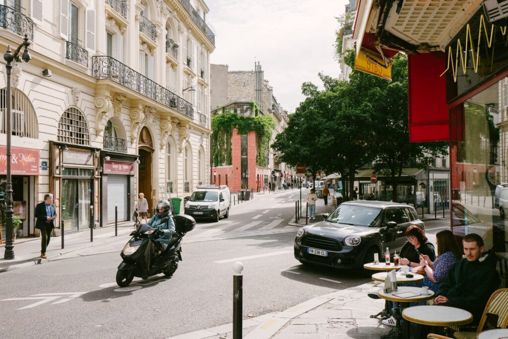 A Paris street scene. On the right people sit outside a cafe. A biker drives past. In the distance a building has bushes growing off it.