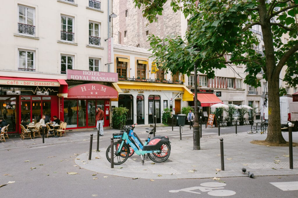 A colourful Parisian street scene. There are two blue dolt bikes. Behind is a white cafe with yellow accents. There is a big green tree and resturants with red ornings.