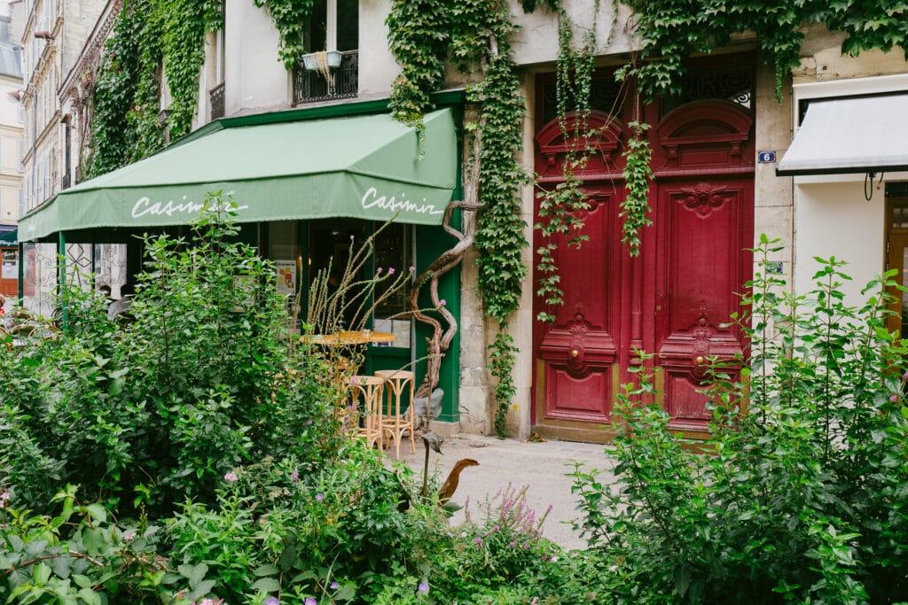 A cafe in Paris with a red door and green orning. The area is covered in plants.