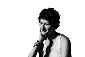 A person with short, curly hair, wearing a sleeveless top, rests their chin on their hand while looking off to the side against a white background.