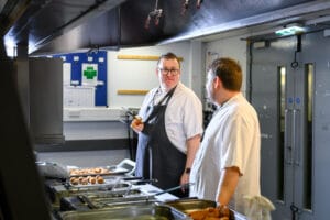 Two chefs are standing in a kitchen, engaged in conversation. One chef, wearing an apron and holding kitchen tongs, is looking at the other chef who is wearing a white shirt. Various food items are in trays in front of them, and there is a bulletin board in the background with papers attached.