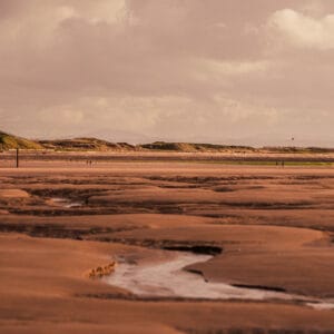 A wide view of a sandy beach at low tide, featuring various patterns in the sand and a small stream flowing through. In the background, green hills and distant mountains are visible under a cloudy sky. A few people can be seen walking on the beach.