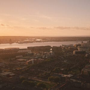 Aerial view of a city at sunset, with a river in the background and buildings, both residential and commercial, in the foreground. The sky is illuminated with warm orange and yellow hues.