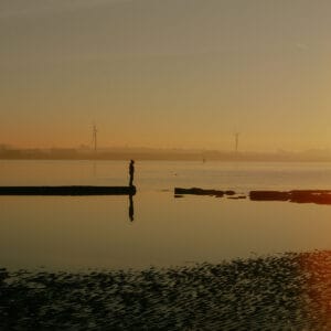 A person stands on a rocky outcrop overlooking a calm body of water at sunrise, with reflections in the water and wind turbines visible in the background against a hazy sky.