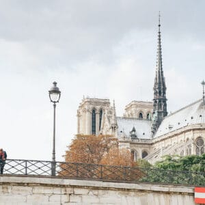 A couple is leaning over a railing by a river, with a view of Notre-Dame Cathedral in the background. A lamppost is nearby, and autumn foliage is visible around the cathedral.