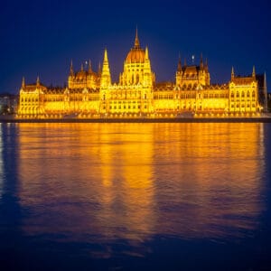 The illuminated Hungarian Parliament building reflected in the Danube River at dusk, with a deep blue sky in the background.