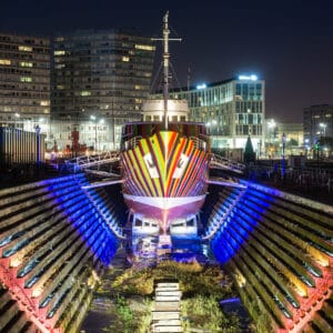 A brightly painted ship sits at night in a dry dock, surrounded by illuminated modern buildings and pathways. The dock features patterned stone steps with colourful lighting, enhancing the urban ambiance.