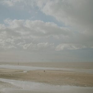 A person walking their dog on a sandy beach, with gentle waves and a distant horizon featuring wind turbines, under an overcast sky with scattered clouds.