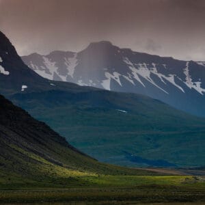 A dramatic landscape featuring steep, rugged mountains with snow-capped peaks under a moody, cloudy sky. The foreground shows sloping green hills with patches of light illuminating the grass, creating a stark contrast with the darker rocky slopes.