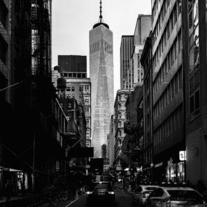 A black-and-white photograph of a narrow street flanked by tall buildings, leading towards a skyscraper in the distance. The skyscraper features a spire at the top, and the scene captures vehicles and pedestrians on the road, along with various storefronts.