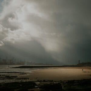A dramatic beach scene under moody storm clouds, with sunlight breaking through. A solitary figure walks along the wet sandy shore, while distant city skyline and buildings are visible across the water. The foreground features rocky textures and water reflections.