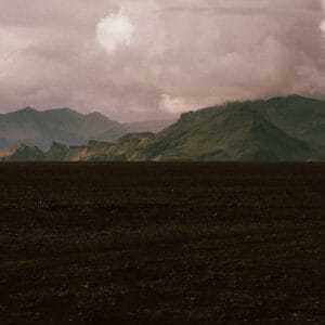 A dramatic landscape featuring a vast, dark, barren terrain with distant mountains under a cloudy sky. The mountains display a mix of green and rocky textures, while the foreground is predominantly black earth.