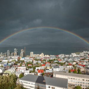 A wide view of a cityscape featuring a vibrant rainbow arching over buildings, with dark clouds overhead and the sea in the background. The scene shows a mix of colourful rooftops and contemporary architecture amidst greenery.