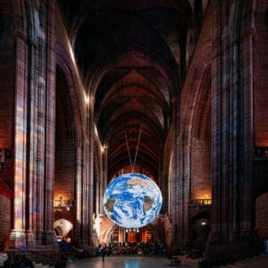 A large globe of Earth suspended from the ceiling inside a grand, dimly lit cathedral, with tall arches and stone walls. The scene is filled with spectators seated below, illuminated by soft lights.