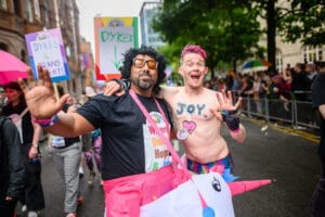 Two men embrace and smile for the camera at Pride. One is a trans man with the word Joy written on their chest with a purple heart beneath it. The other is riding a unicorn.