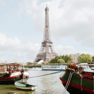A view of the Eiffel Tower in Paris, framed by boats moored along the riverbank, under a blue sky dotted with clouds.
