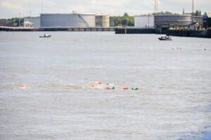 A pod of swimmers swim ahead of the main group of swimmers. There are safety boats present as they pass an industrial area.