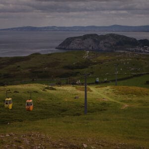 A landscape view featuring green hills with cable cars in yellow and orange descending, overlooking a calm sea and a distant coastal area under a cloudy sky.