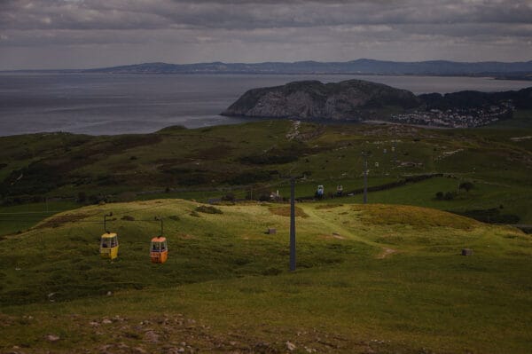 A landscape view featuring green hills with cable cars in yellow and orange descending, overlooking a calm sea and a distant coastal area under a cloudy sky.