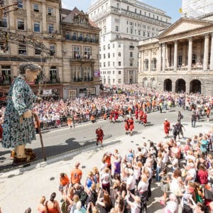 A large crowd gathers in a city square to watch a giant puppet resembling an elderly woman, dressed in a patterned gown and holding a cane. The scene features various performers in red costumes, with historic buildings in the background and a bright blue sky overhead.