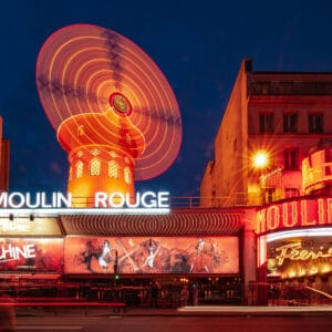 Illuminated facade of the Moulin Rouge cabaret in Paris at dusk, featuring a spinning red windmill and vibrant neon signs.